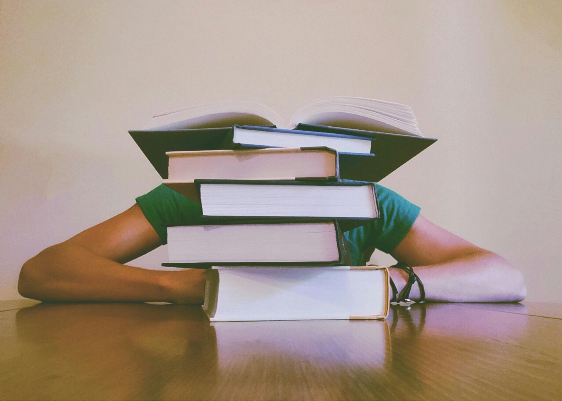 A person in a green shirt sits at a wooden table with a large stack of books covering their head and face.