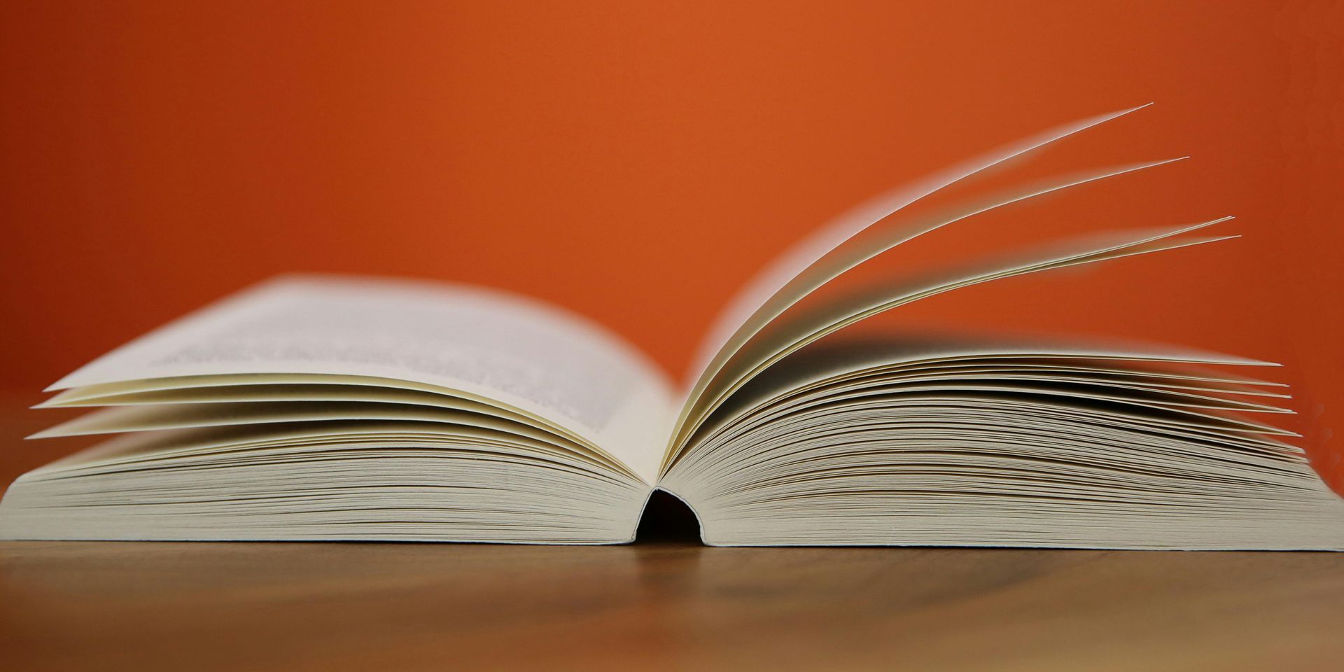An open book with visible pages rests on a wooden surface against a solid orange background.
