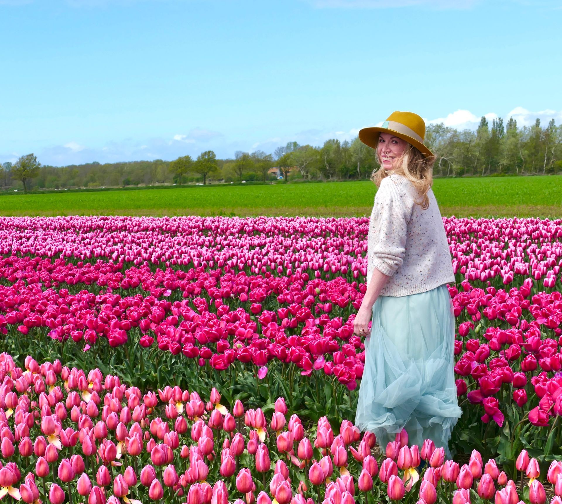 A smiling person in a hat, light top, and long skirt stands in a vast, sunny field of bright pink and purple tulips.