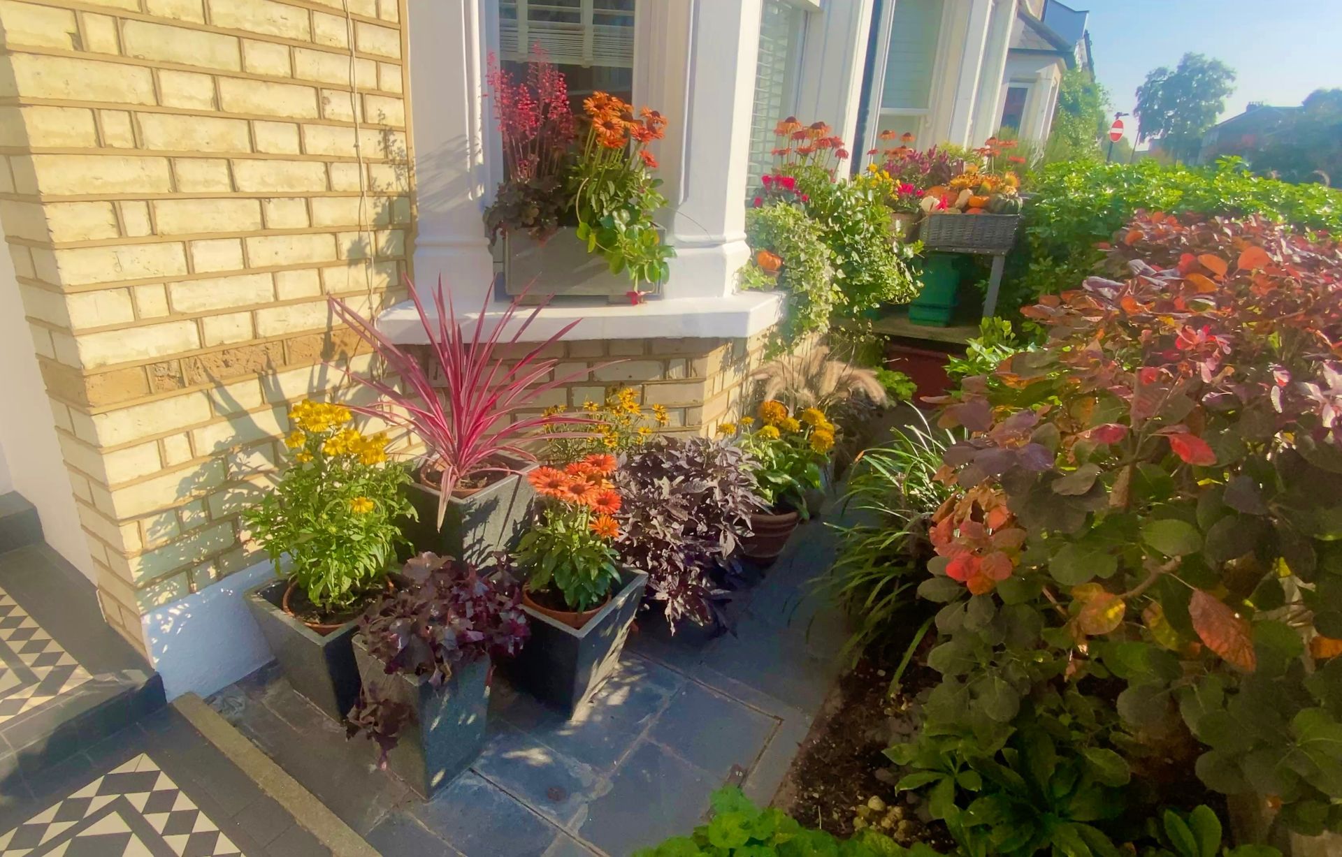 Yellow brick house facade with planters and pots filled with colorful flowers and lush plants on a tiled entryway.