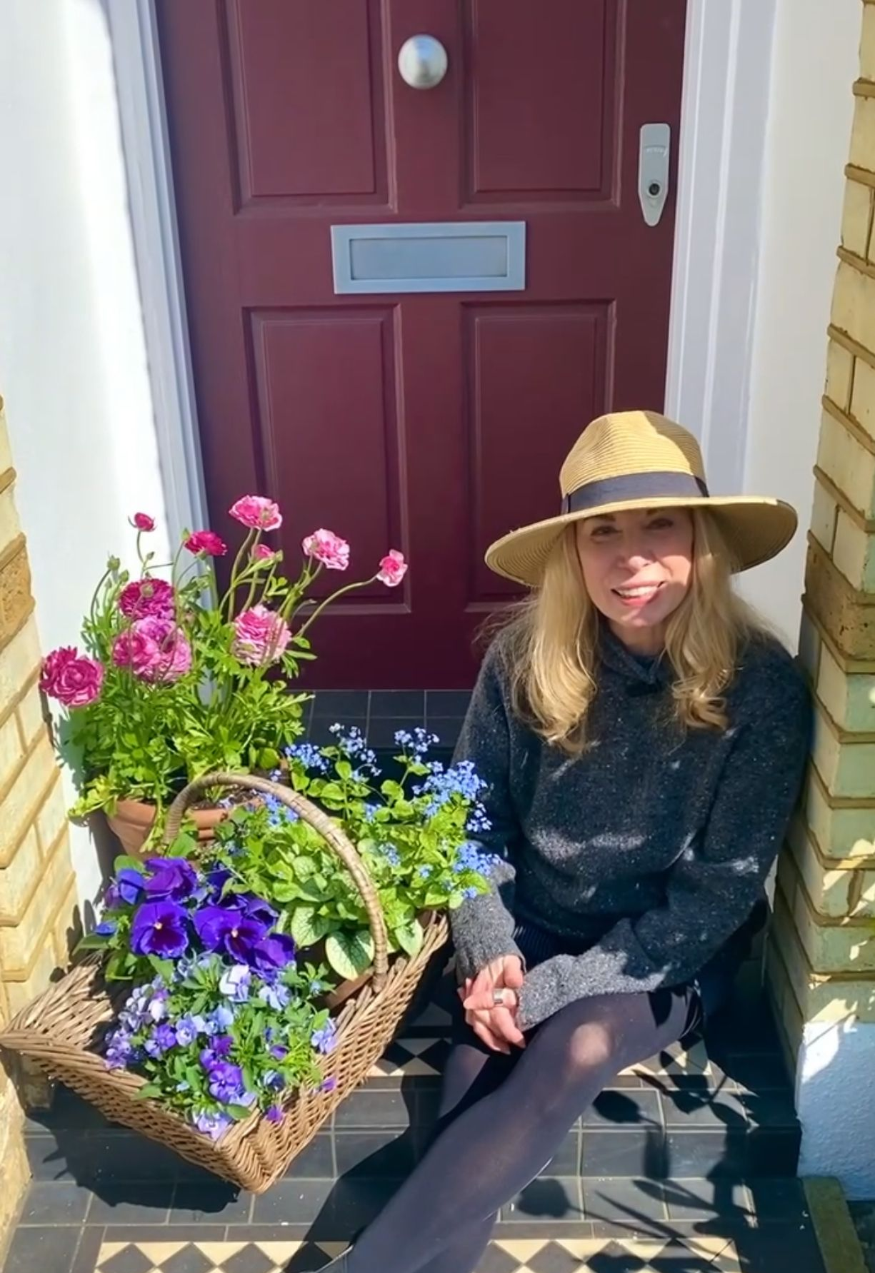 A person in a sun hat sits by a red front door beside a basket of purple and blue flowers on a tiled step.