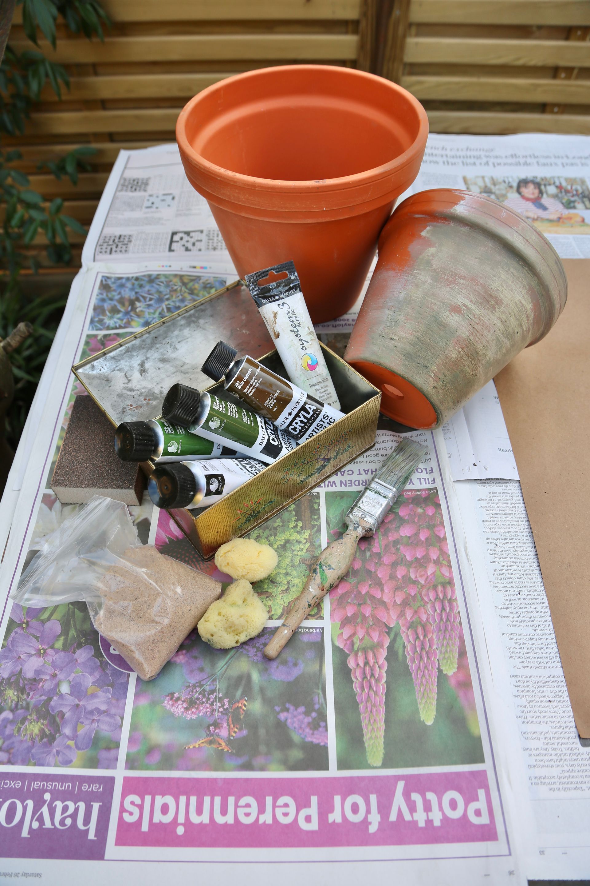 Terracotta pots, acrylic paints, brushes, and sponges arranged on newspaper, ready for a craft project.