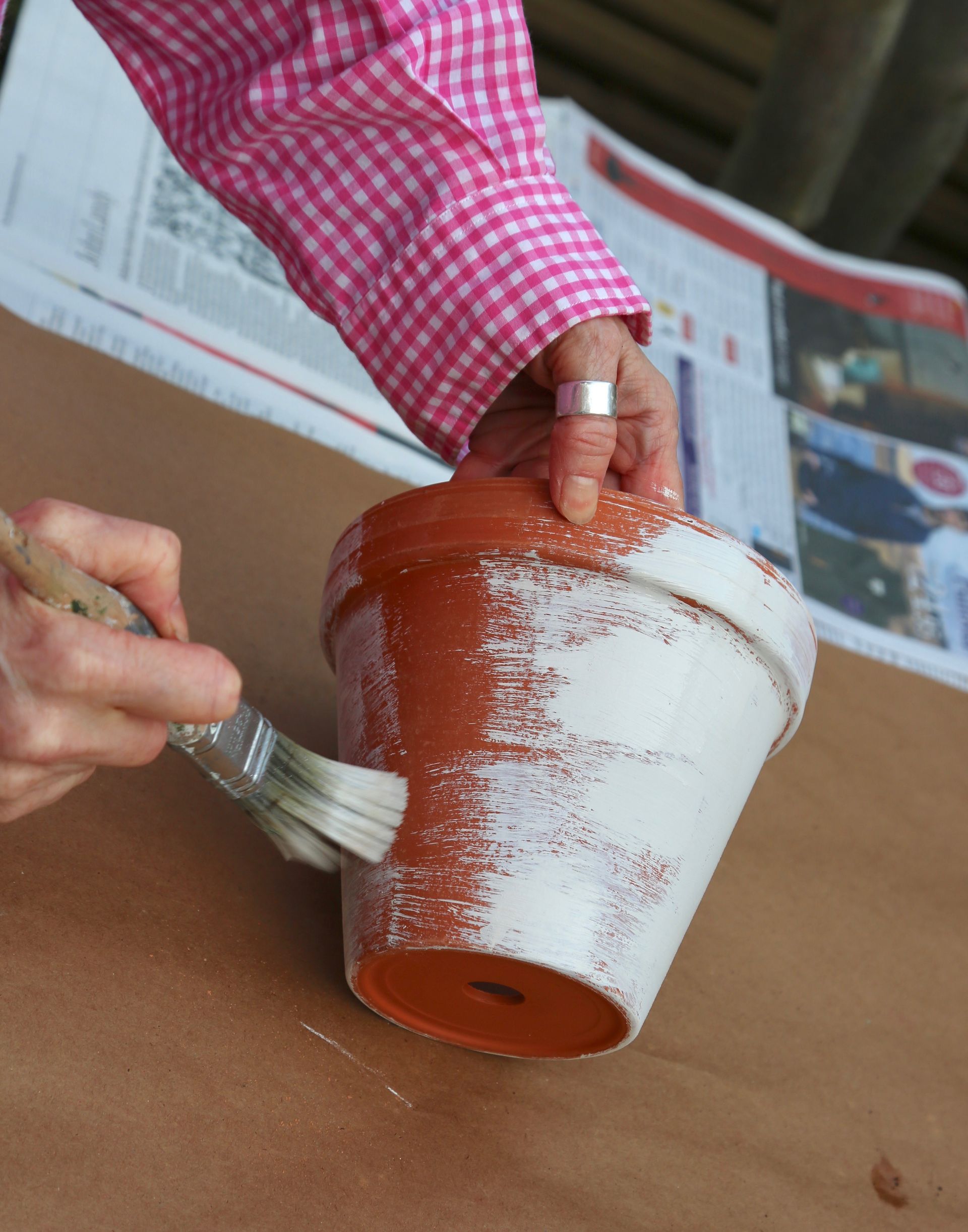 A person wearing a pink checkered shirt uses a paintbrush to apply white paint to a terracotta clay pot.