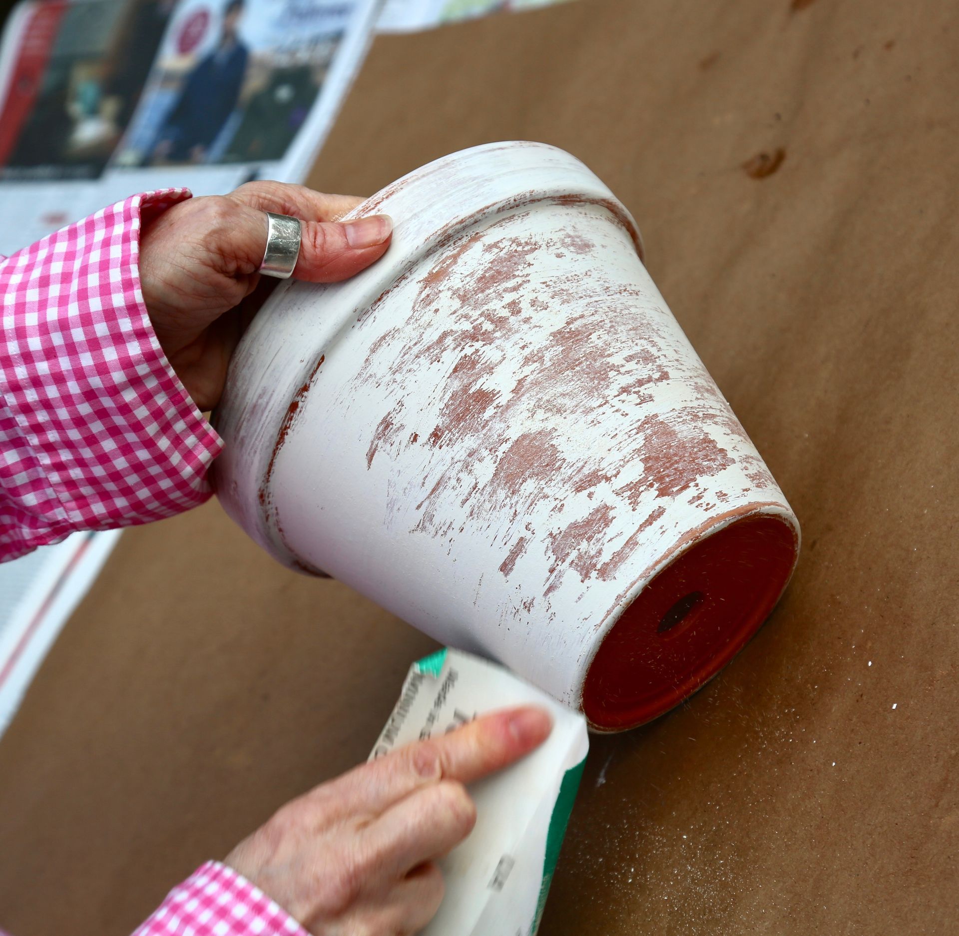 A person wearing a pink checkered shirt uses a sanding block to distress a white-painted terracotta flower pot.