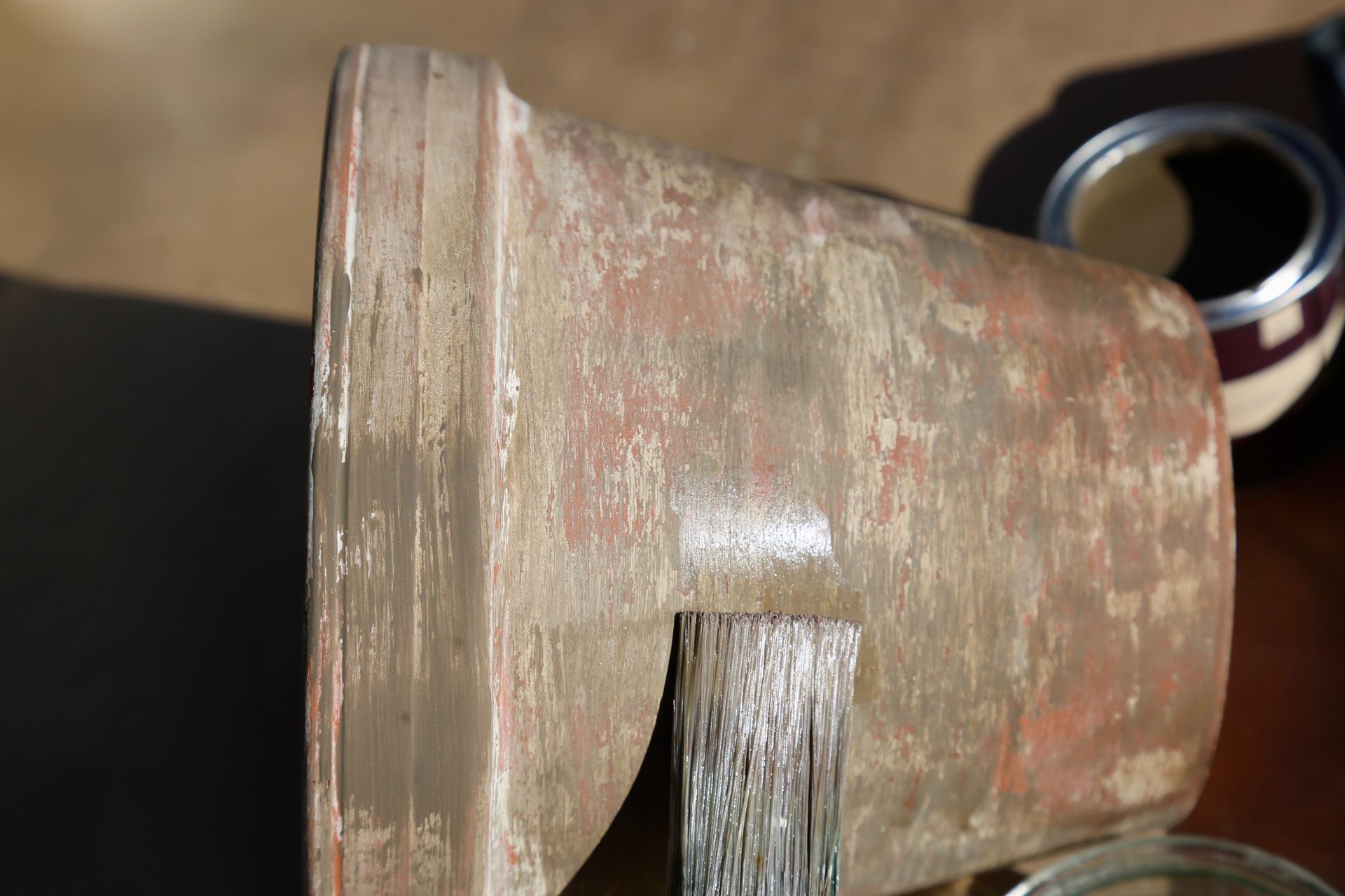 A paintbrush applies a coat of neutral-toned paint to the textured exterior of a terra-cotta flower pot.