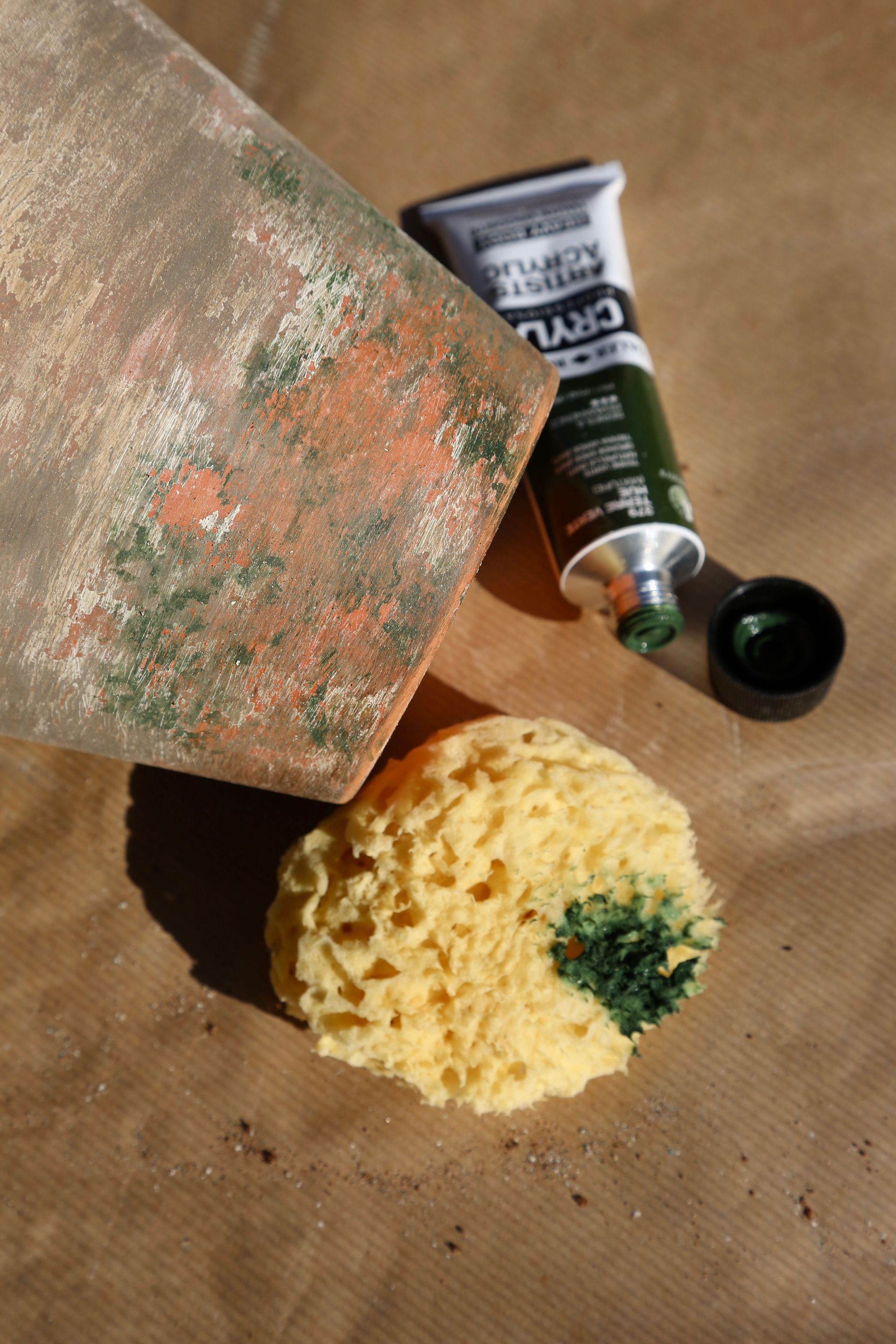 A terracotta pot being painted with dark green acrylic paint using a natural sponge on a brown paper surface.