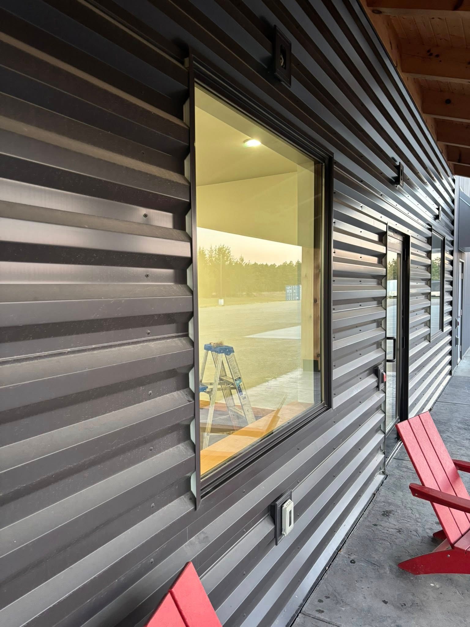 Exterior of a building with dark gray corrugated metal siding, large window, red chairs.