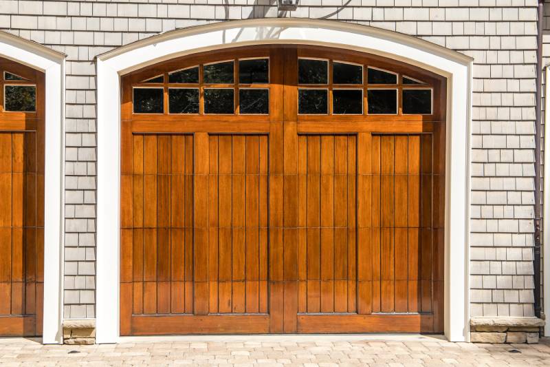 Arched wooden garage door with glass window panes set in a white brick facade, showcasing a classic design.