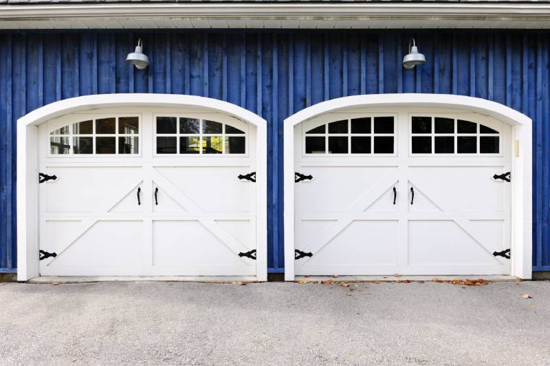Dual white garage doors with arched windows and black hardware on a blue facade, offering a traditional aesthetic.