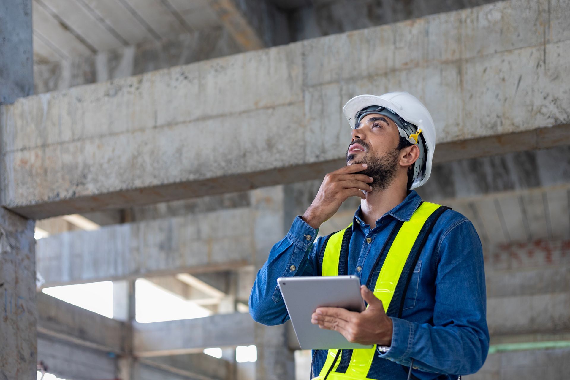 A construction worker is looking up at the ceiling while holding a tablet.