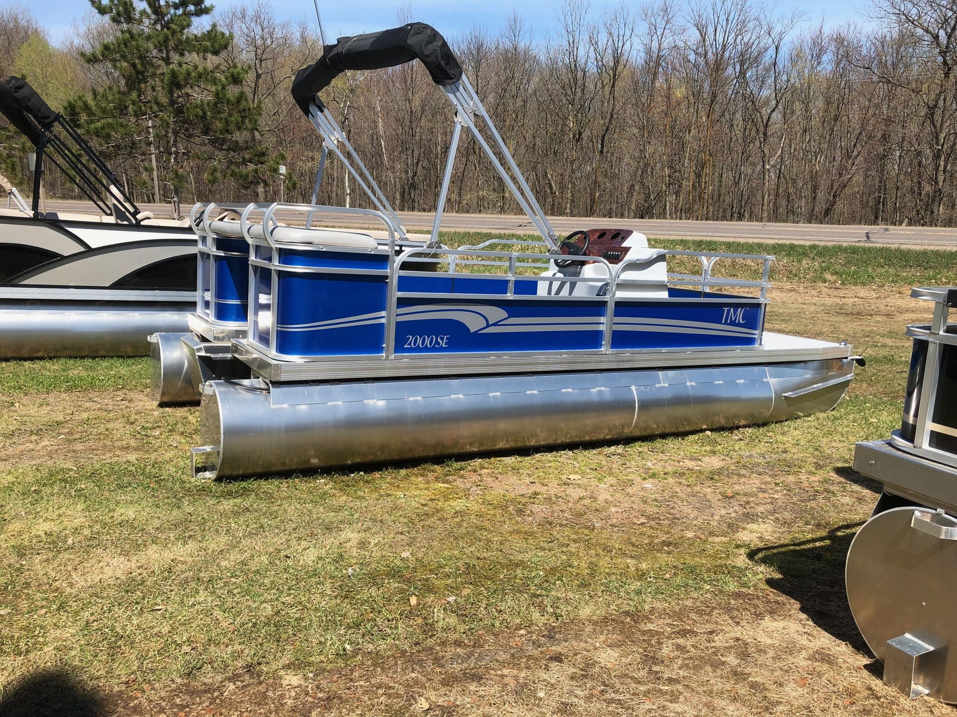 Blue and silver pontoon boat with a black canopy on grassy ground.