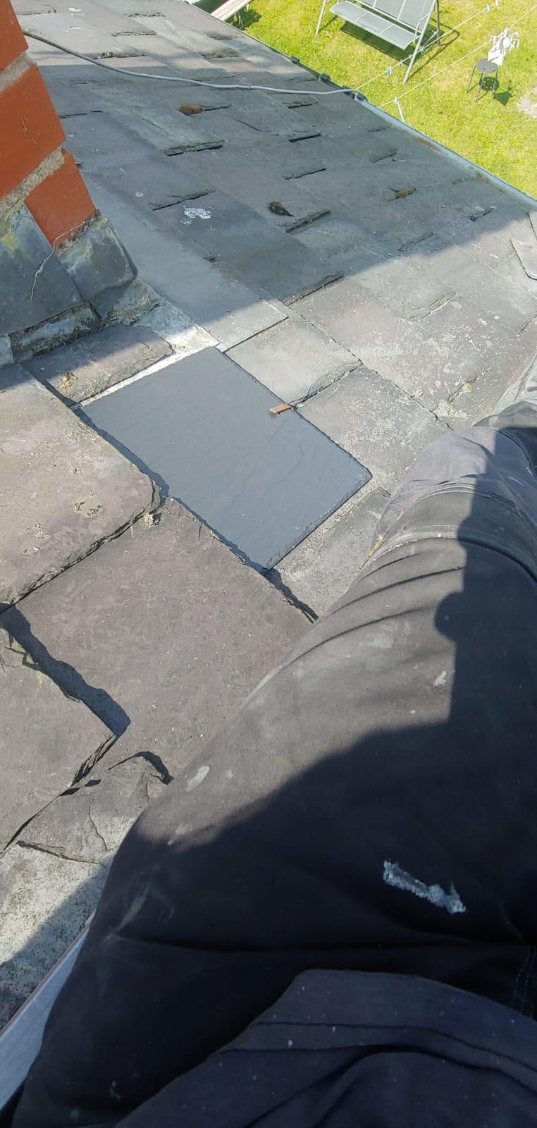 Person sitting on a roof, next to a brick chimney, looking down at a dark square panel set within the roof tiles.