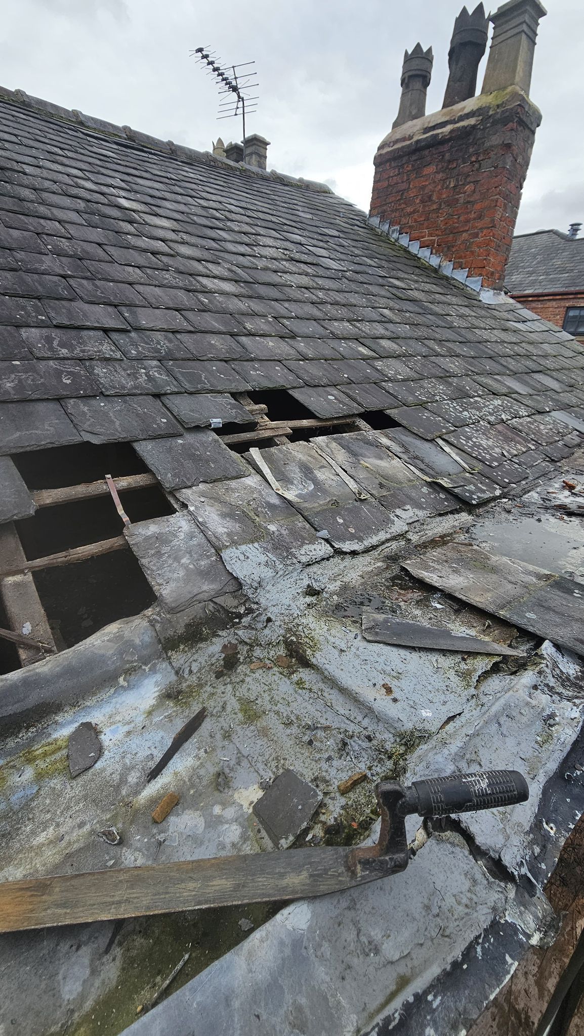 Damaged roof with missing tiles, exposing wooden structure. Chimneys and an antenna are in the background.