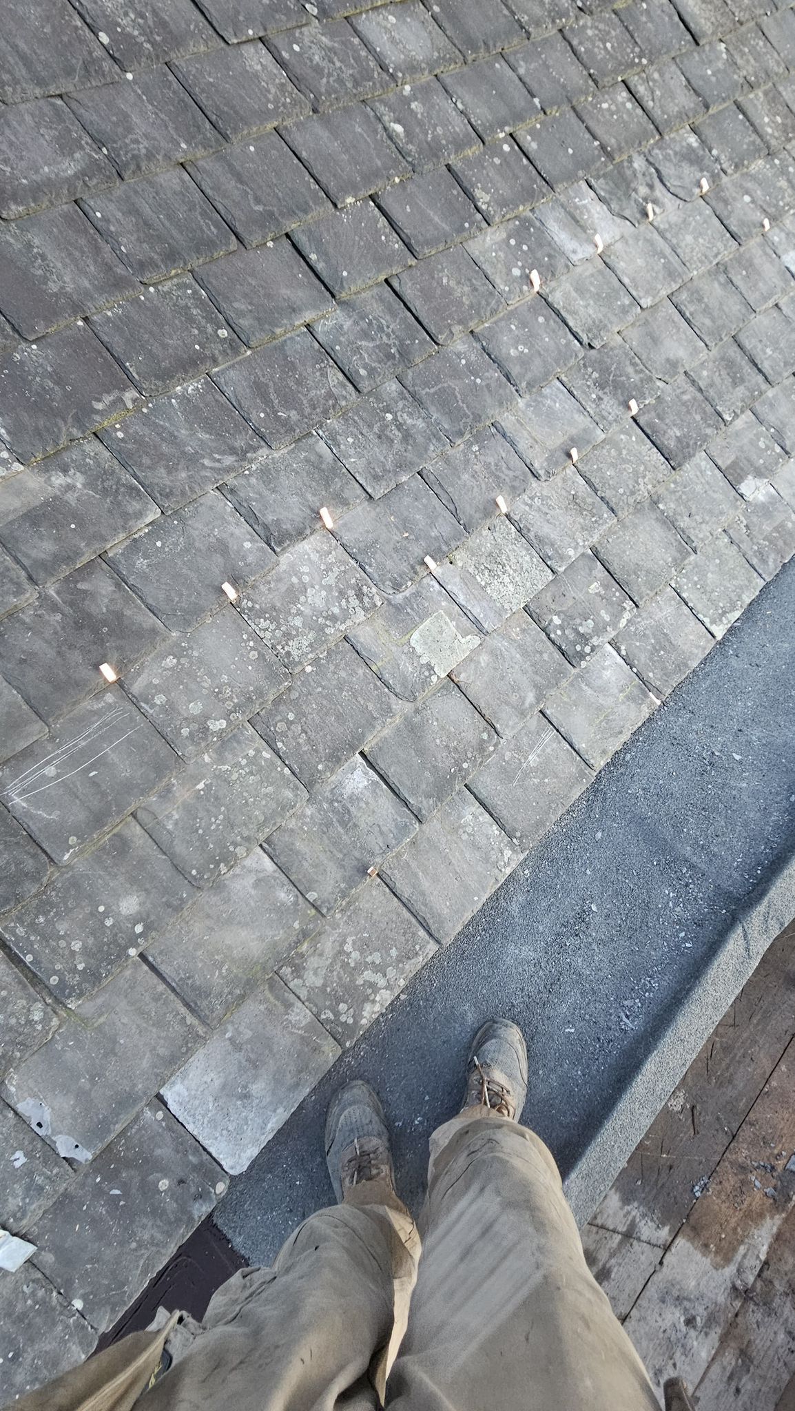 Person's legs and feet standing on a gray asphalt shingle roof.