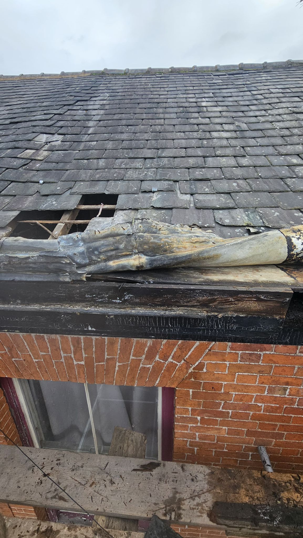 Damaged roof with missing shingles and burnt wood, above a brick building with a window.