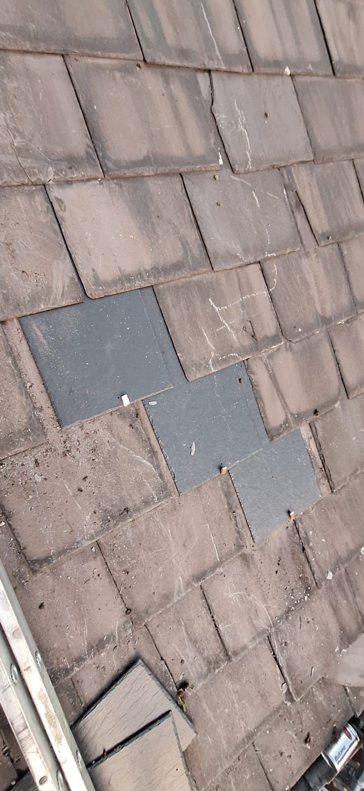 Close-up of a roof with several dark gray replacement tiles among the weathered brown tiles.
