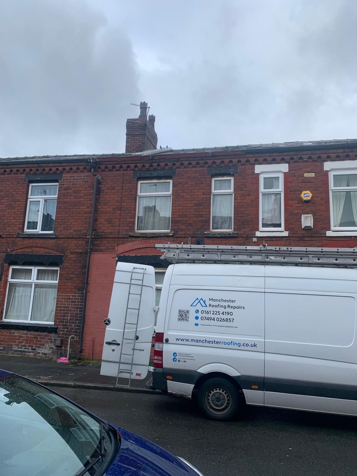 Red brick row houses; Manchester Roofing Repairs' white van parked on street with ladder resting on the roof. Cloudy day.