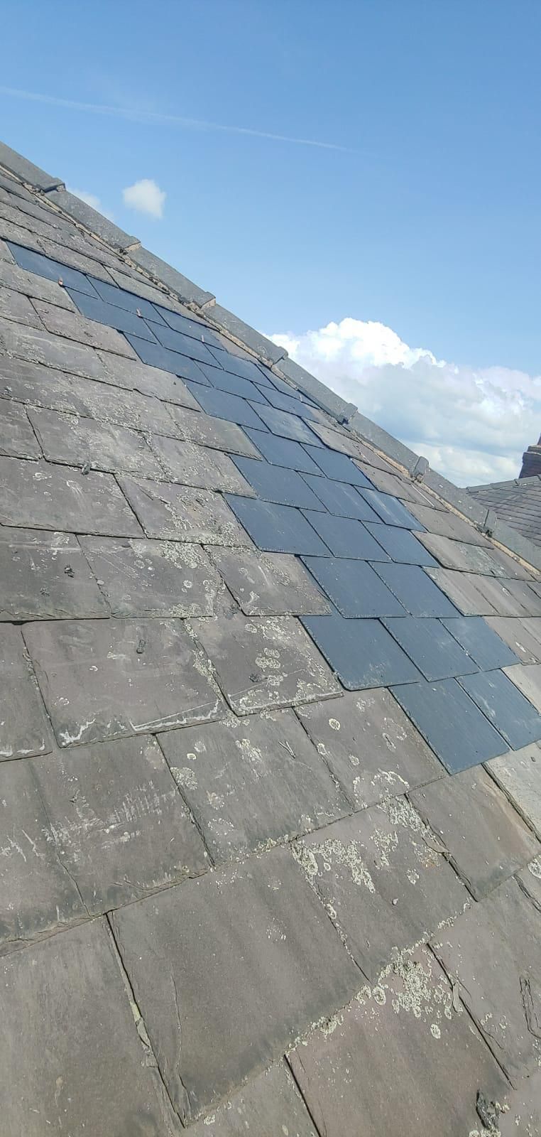 A rooftop with a patch of dark solar panels installed on weathered gray tiles, against a blue sky with fluffy clouds.