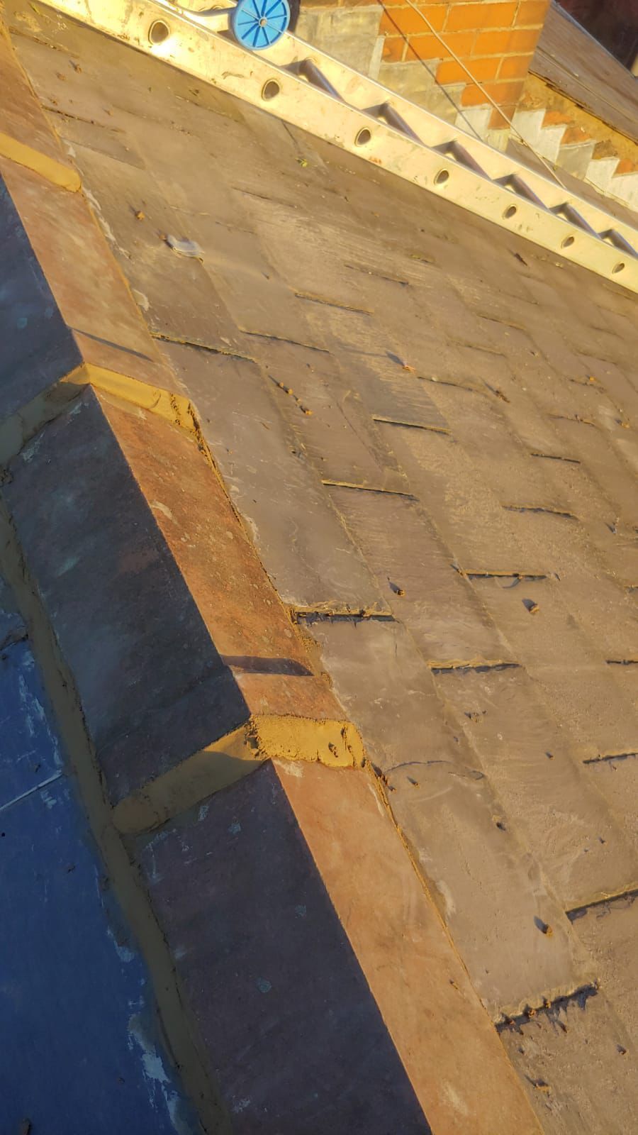 Close-up of a weathered, dark-gray tiled roof with wooden beams, bathed in sunlight. Staircase visible in background.