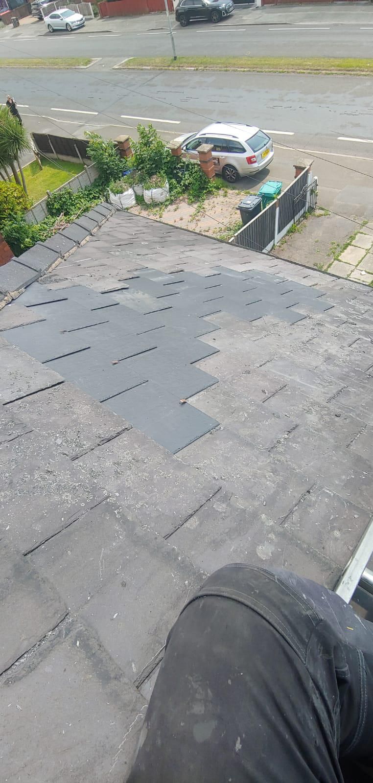 A person kneels on a weathered roof patching asphalt shingles. A street with cars and a white vehicle parked nearby is in the background.