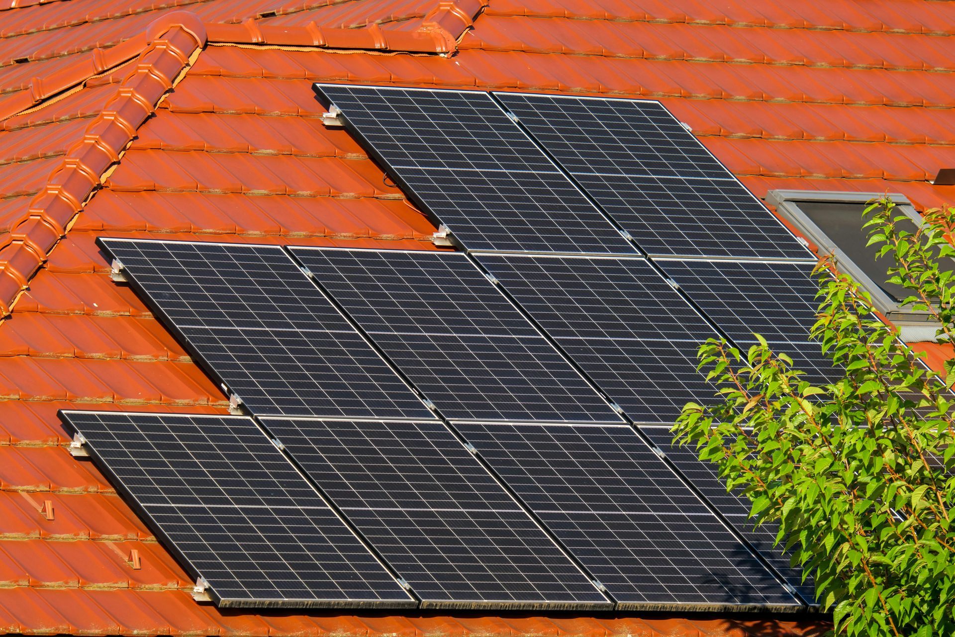 A solar panel array installed on an orange, tiled pitched roof, with a glimpse of green foliage on the right side.