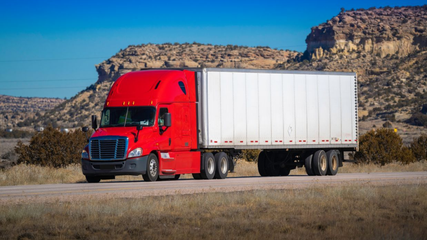 Red semi-truck with white trailer on a road in a desert landscape with rocky hills in the background.