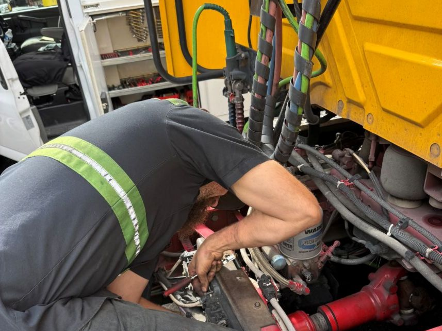 Mechanic working under the hood of a yellow truck, wearing a gray shirt with a green stripe.