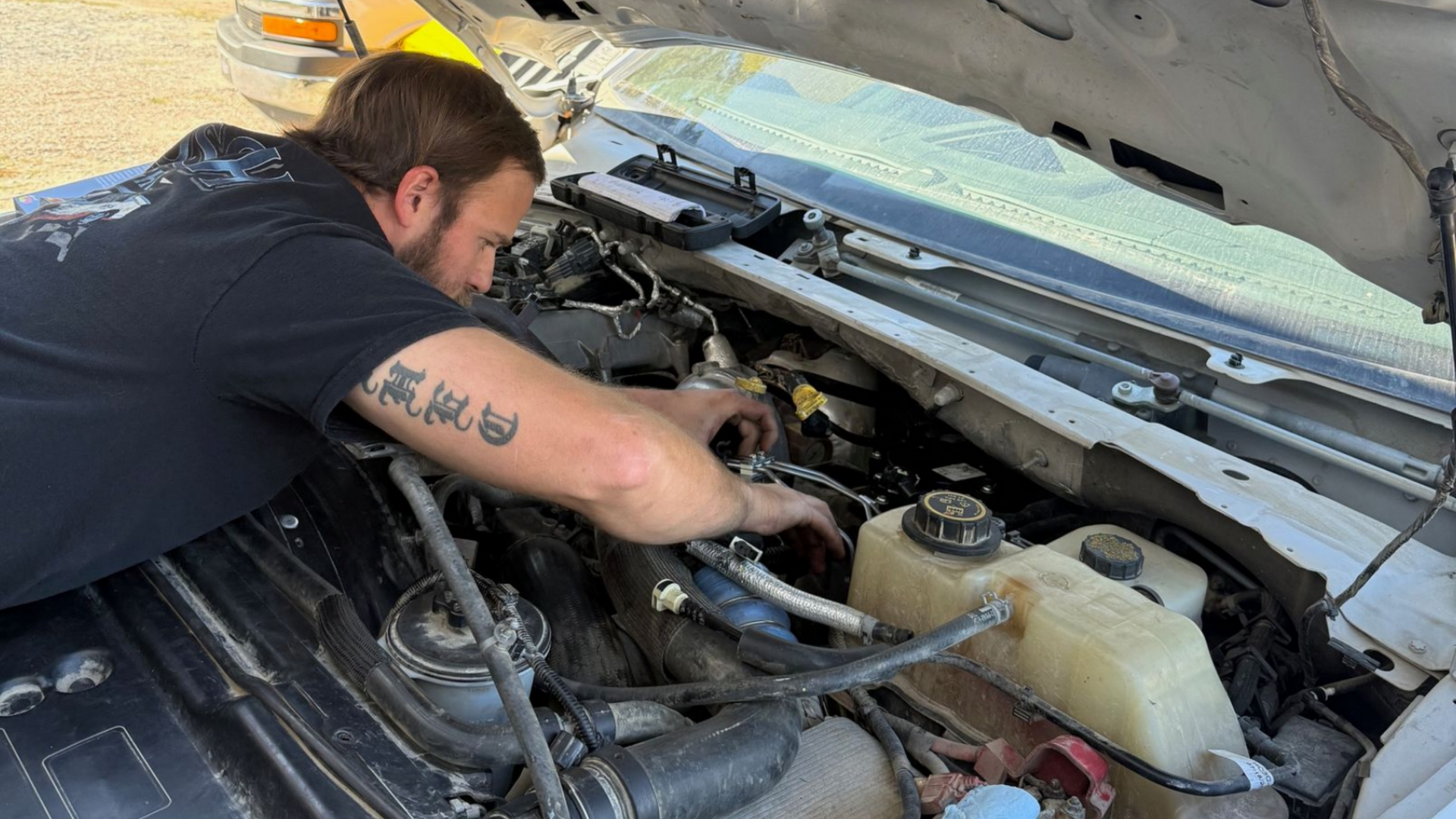 Man working on a car engine, outdoors. He wears a black shirt, is focused, and leans over the open hood.