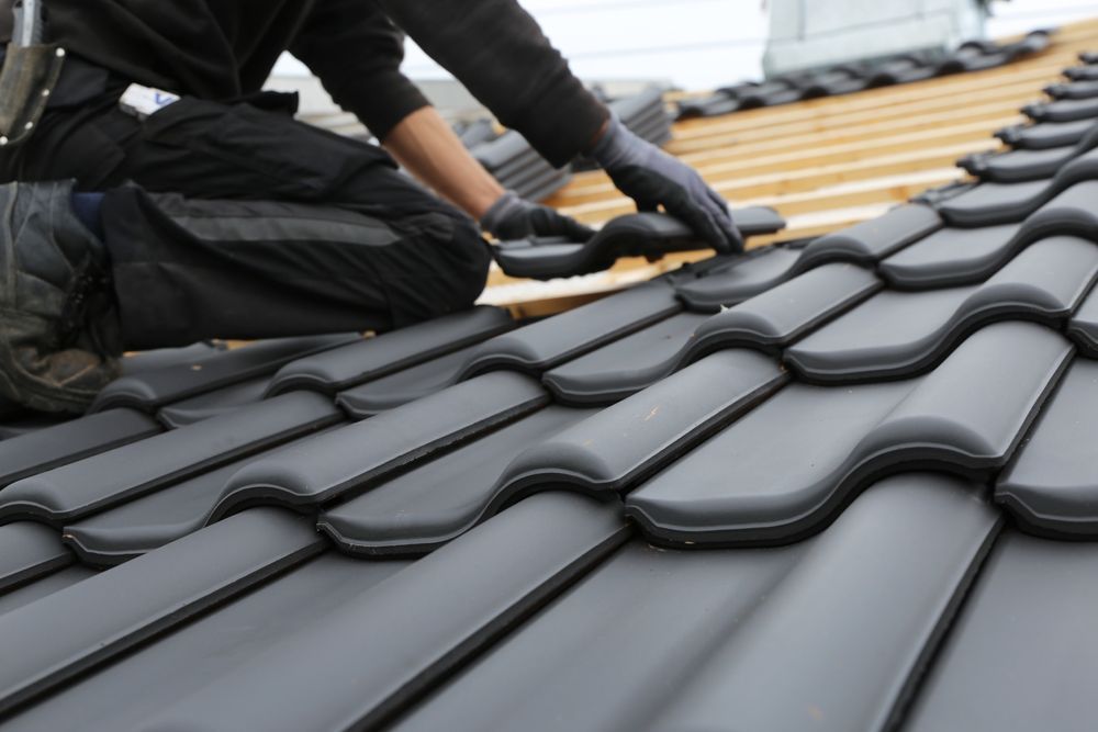 Roofer installing dark gray roof tiles. Rooftop setting, person wearing black pants and gloves.