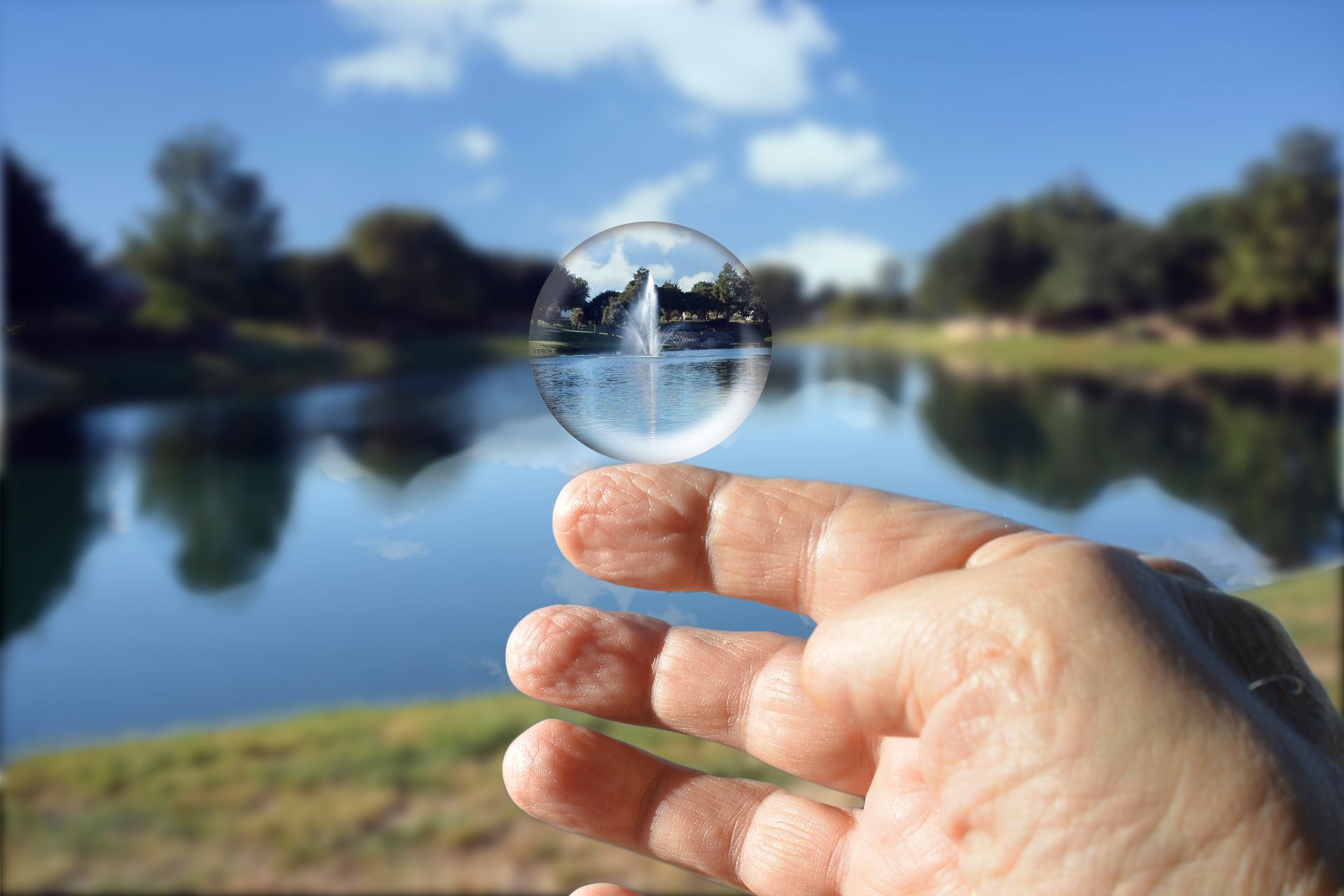 A hand holds a clear lens, which focuses on a fountain in a lake with trees and blue sky.