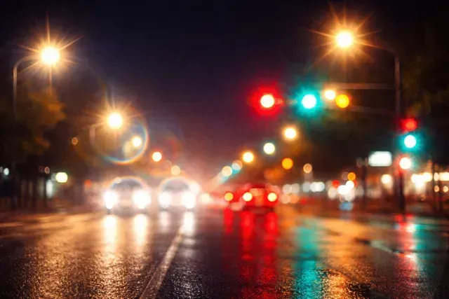 A blurry, out-of-focus night scene of a wet city street with vehicle headlights and traffic lights reflecting on pavement.
