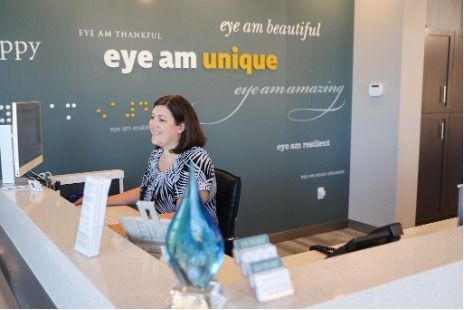 A woman is sitting at the front  desk  at Focal Pointe Eye Care in front of a wall that says eye am unique