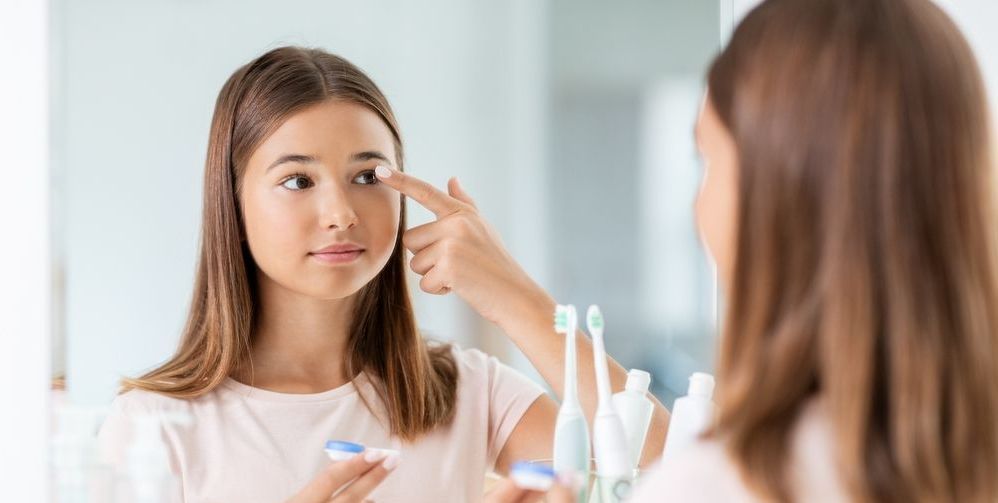 A teenager is applying contact lenses to her eye in front of a mirror.