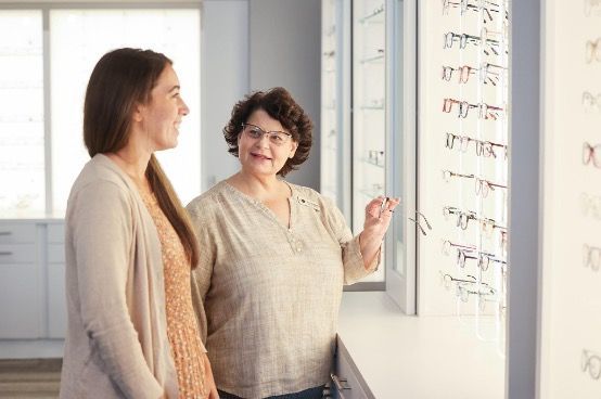 An optical specialist and a patient in front of a wall with glasses on it.