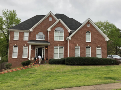 Two-story brick house with white trim, green lawn, and worker near the entrance with hose.