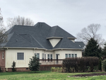Beige house with dark gray shingled roof, brown deck, brick foundation, and leafless trees against a cloudy sky.
