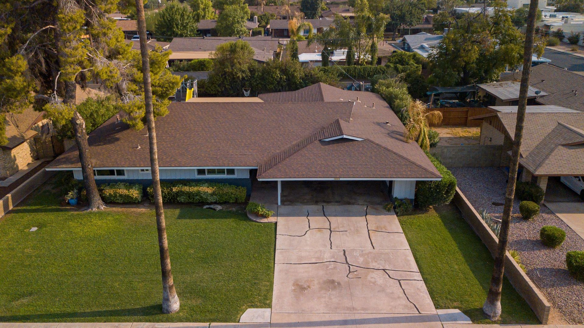 Aerial view of a single-story house with a driveway. Brown roof, green lawn, and trees surround the property.