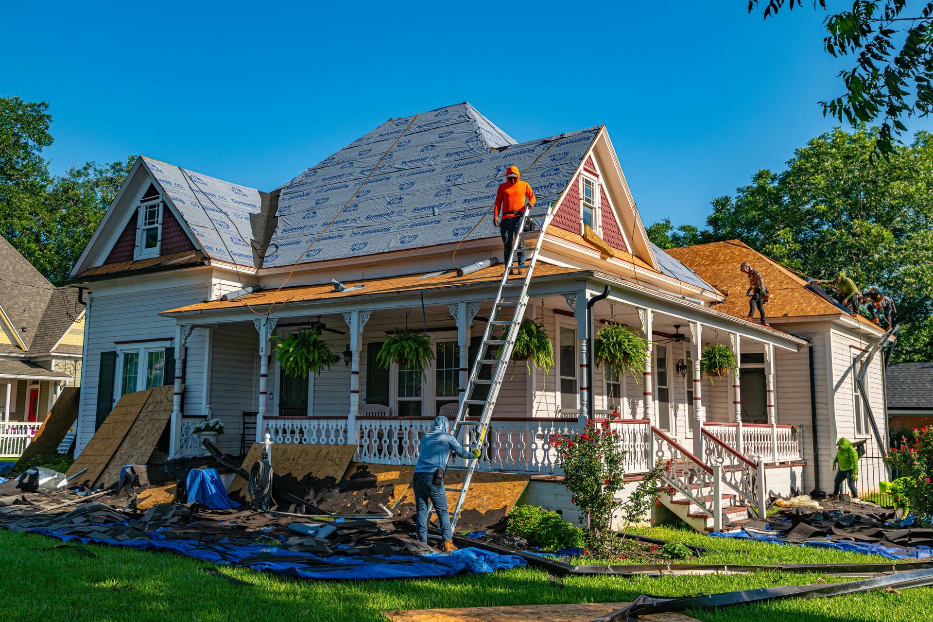 Workers replacing the roof of a two-story white house. One worker on a ladder, two on the roof. Bright sunny day.