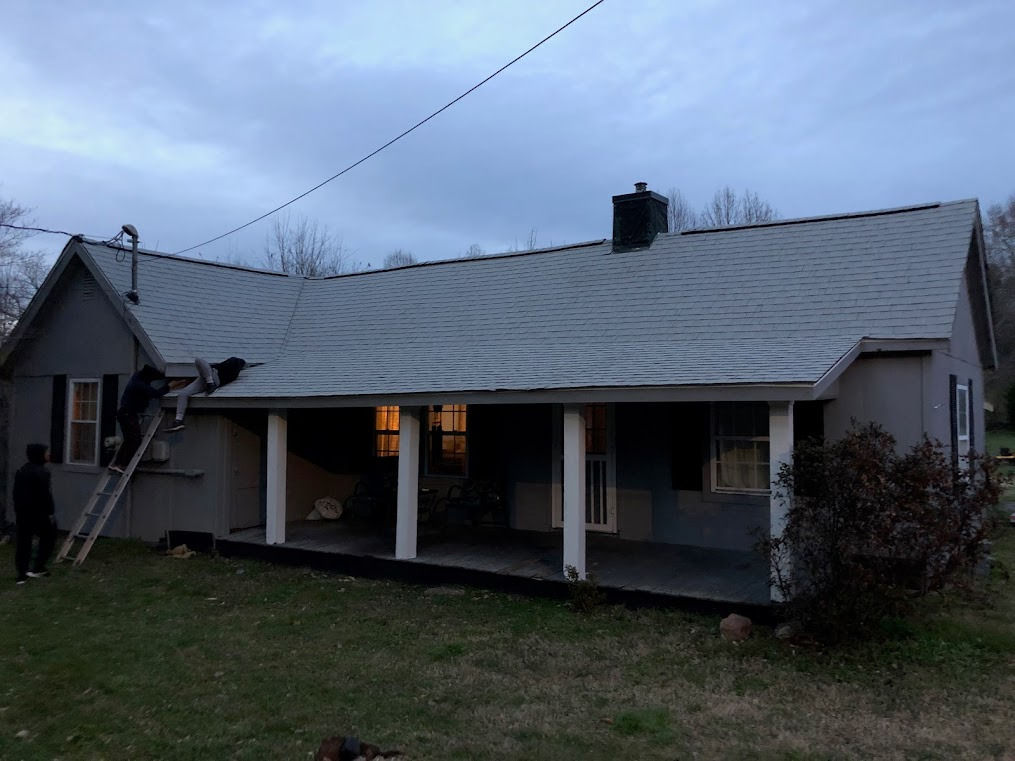 Gray house with porch, workers on a ladder installing shingles under a cloudy sky.