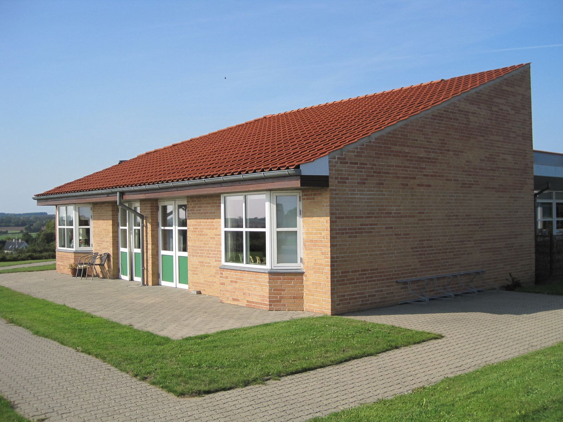 Murstensbygning med rødt tegltag og vinduer, beliggende på et græsareal, en murstenssti og blå himmel.