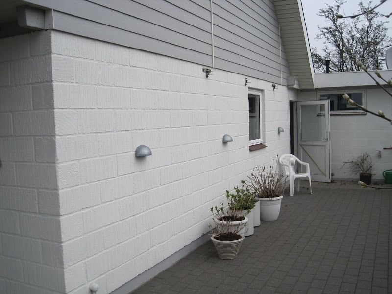 Hvidmalet murstensbygning med grå lister. Plantepotter omkranser en terrasse; dør og vindue er synlige.