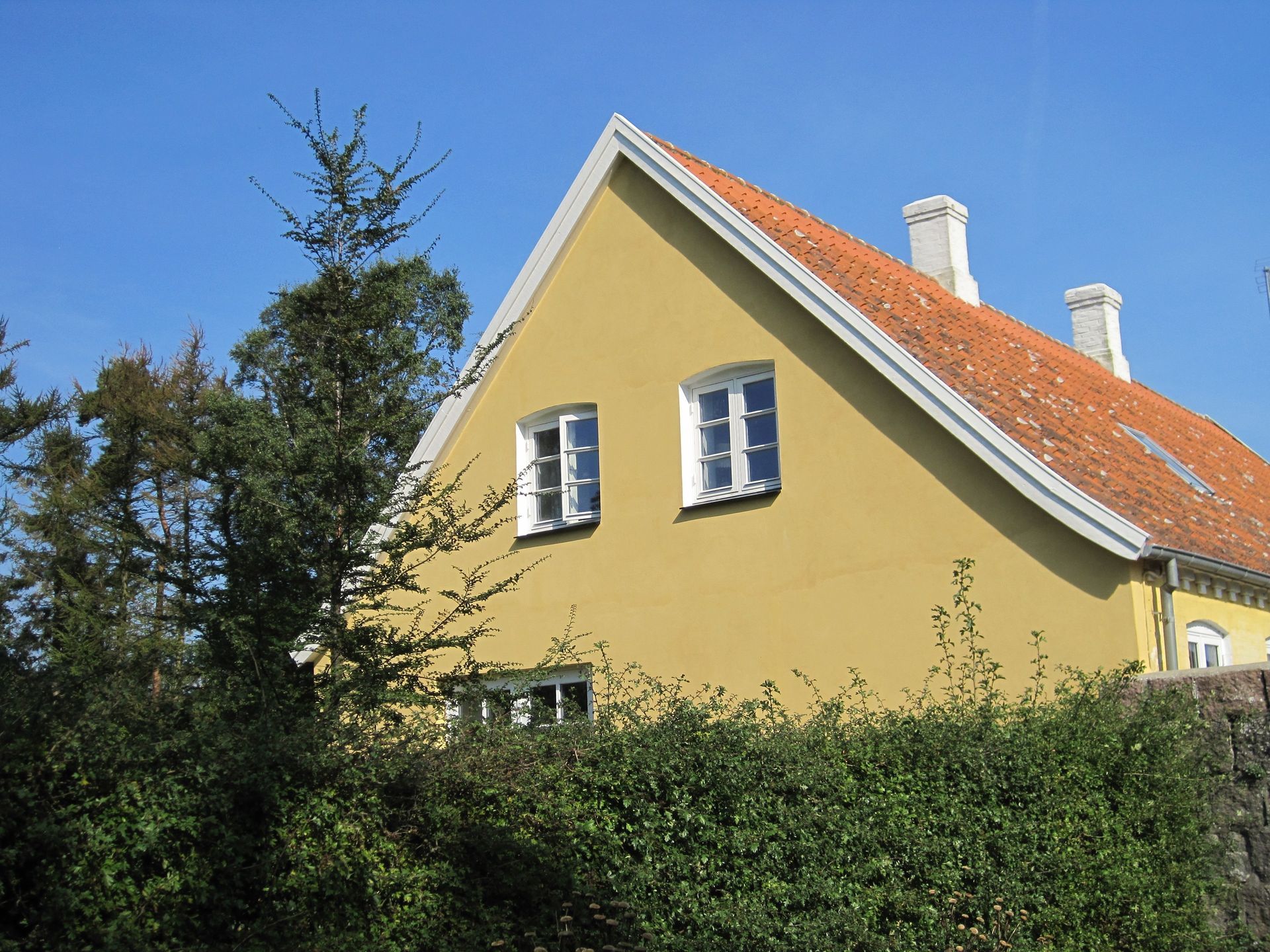 Gult hus med hvide lister, rødt tegltag og to skorstene mod en klar blå himmel.