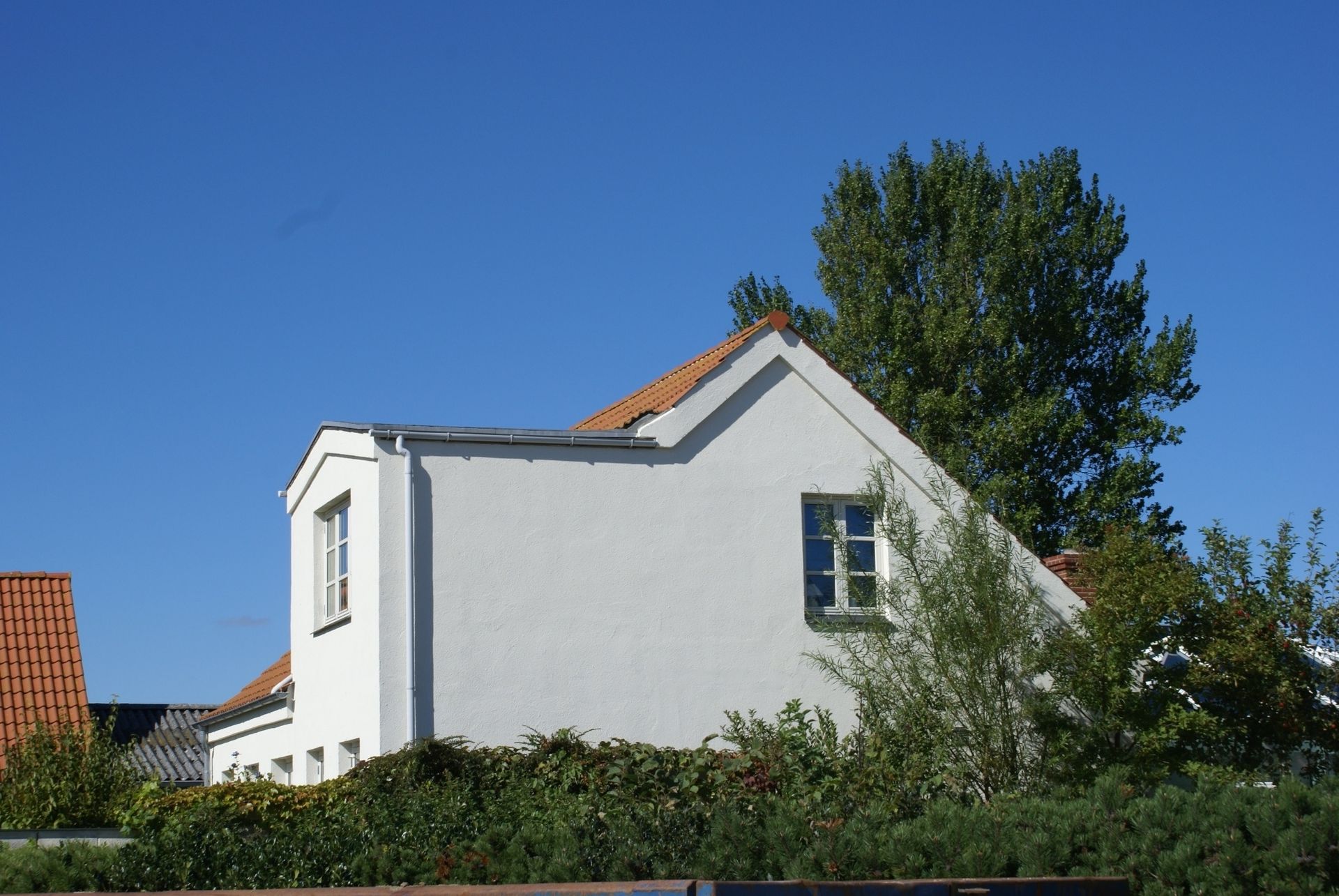 Hvidt hus med rødt tegltag, to vinduer, blå himmel og grønt træ.