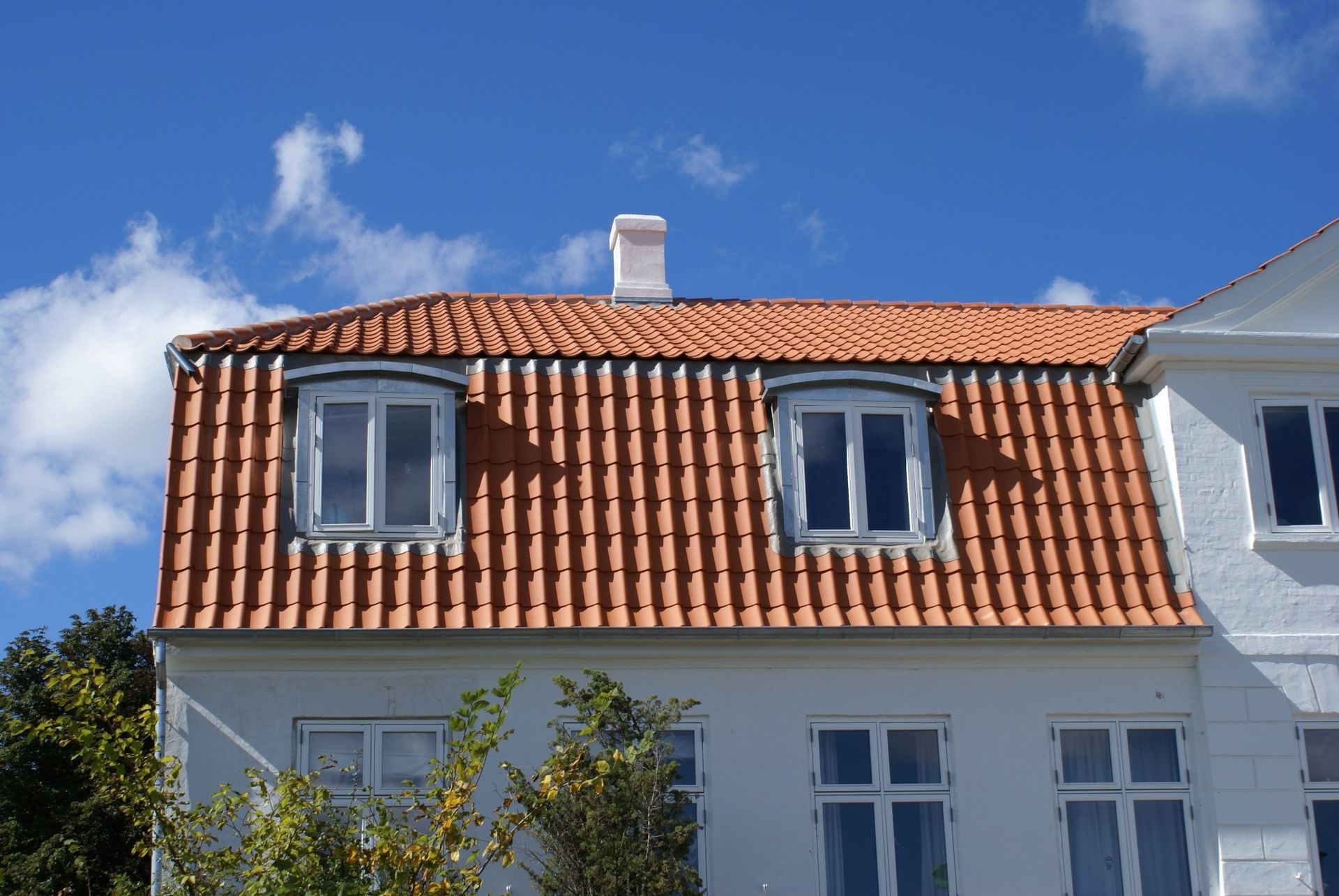 Hvid bygning med orange tegltag og to kvistvinduer mod en blå himmel med skyer.