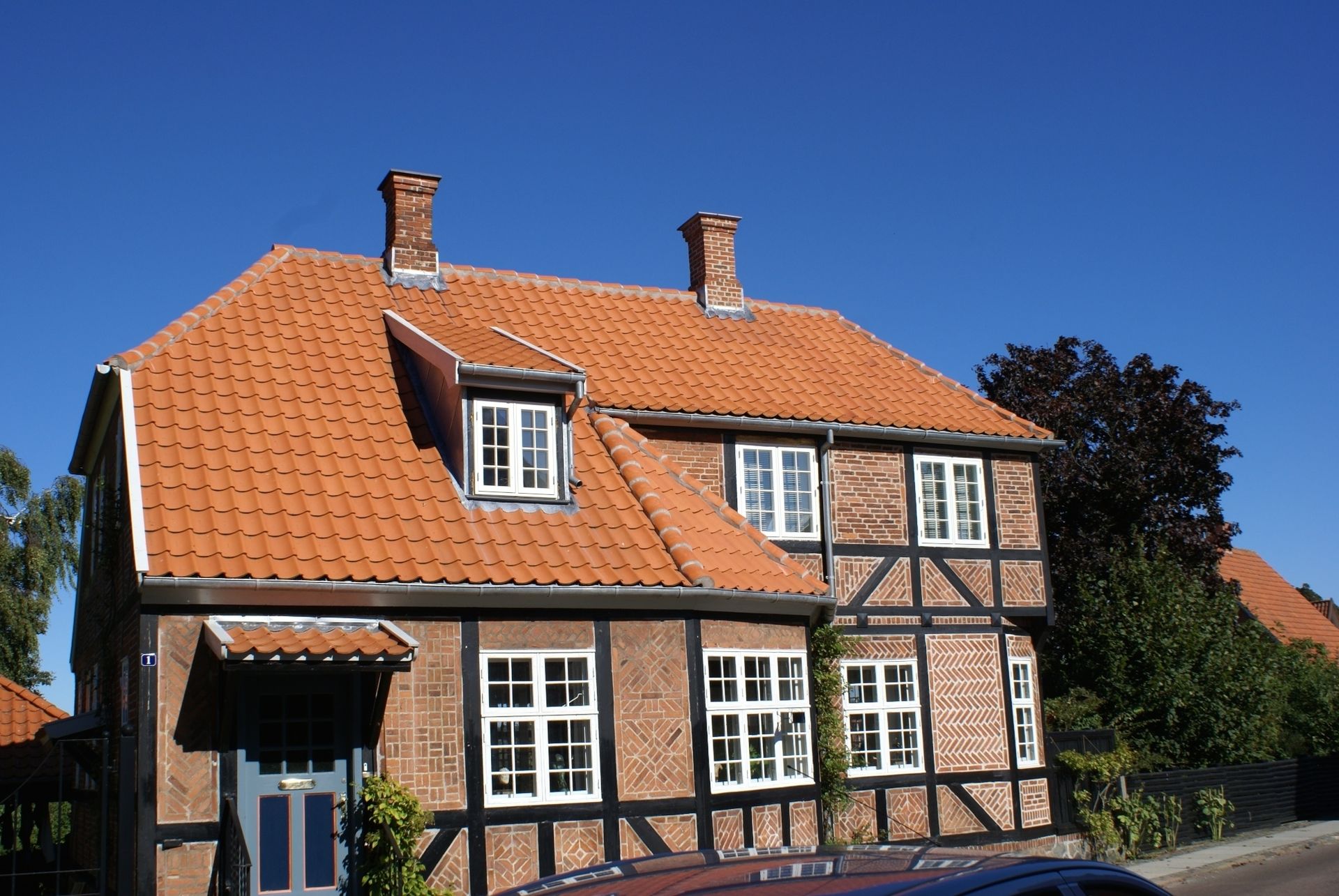 To-etagers murstenshus med rødt tegltag og mørke træbjælker mod en klar blå himmel.