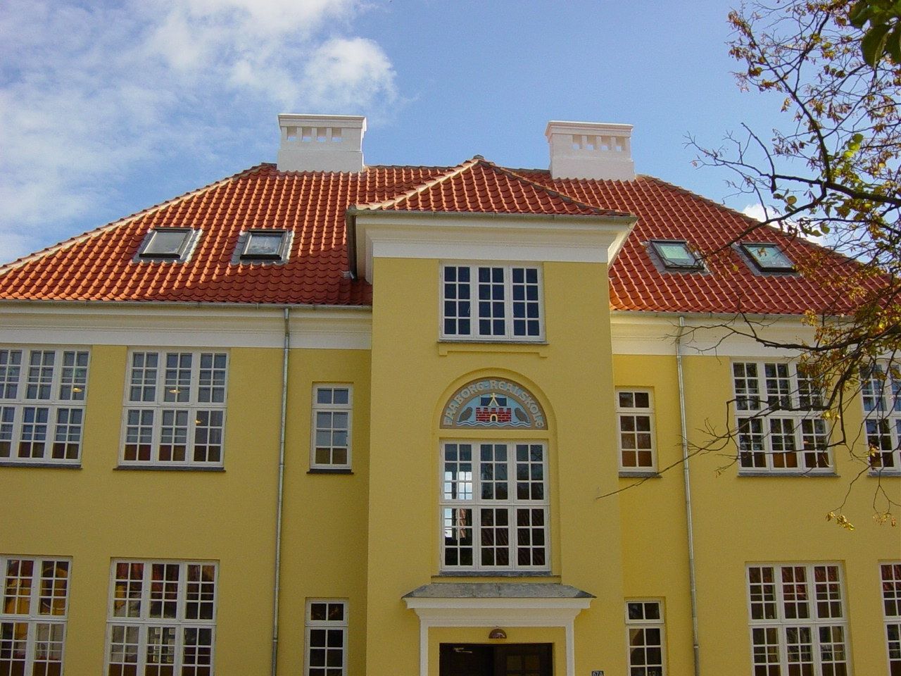 Gul bygning med rødt tegltag, hvide vinduesrammer og blå himmel.