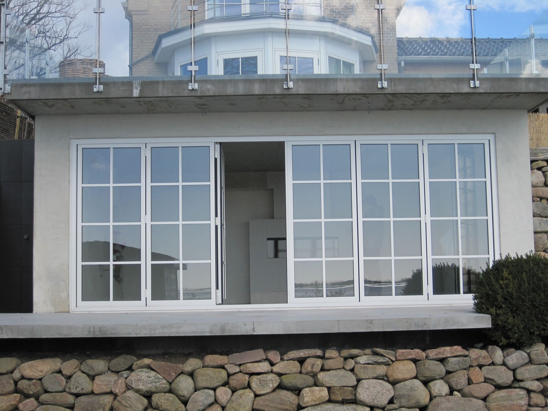 Hvidindrammede vinduer og dør på en betonkonstruktion. Stenmur forneden, glasgelænder foroven.