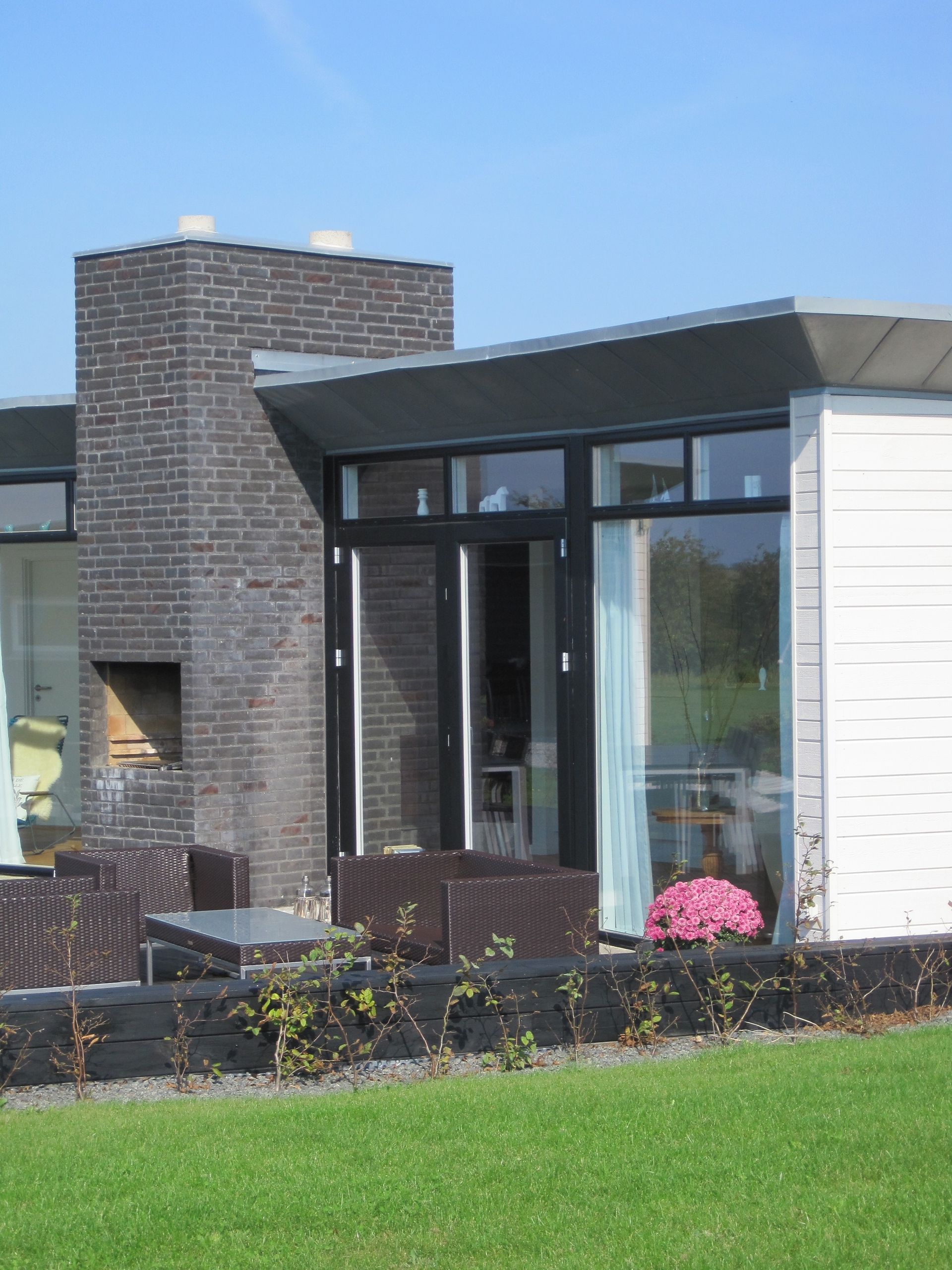 Moderne hus med murstensskorsten, havemøbler, store vinduer og grøn græsplæne.