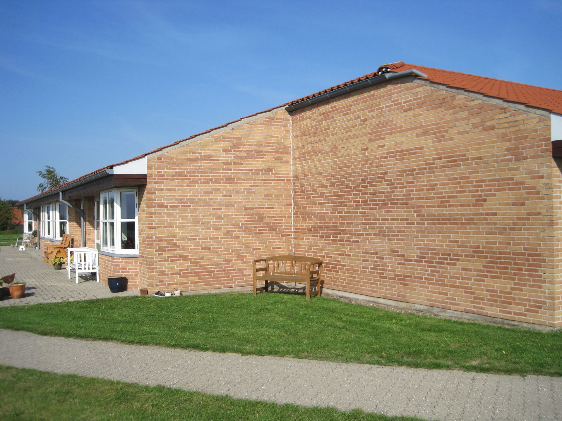Brun murstensbygning med græsplæne, bænk og fortov. Vinduer og blå himmel er også synlige.