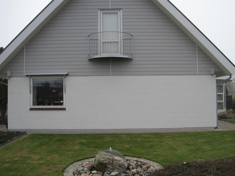 Gråt og hvidt husfacade med en lille balkon og en forhave med et stenfontæne.